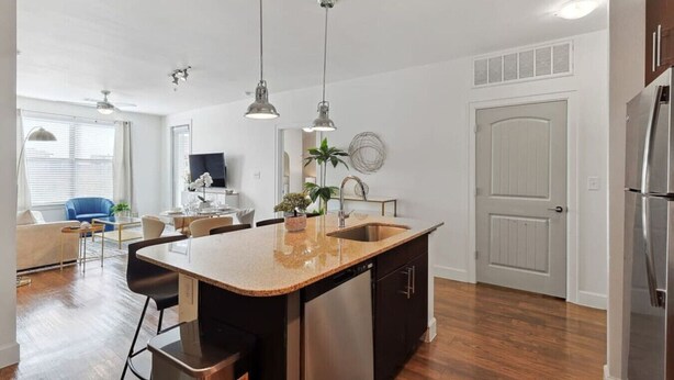 A modern kitchen with a central island, stainless steel appliances, pendant lighting, and a dining area with a blue chair. - Elegant 2B Near Legacy West Wi-fi, Fitness, Pool (Frisco)