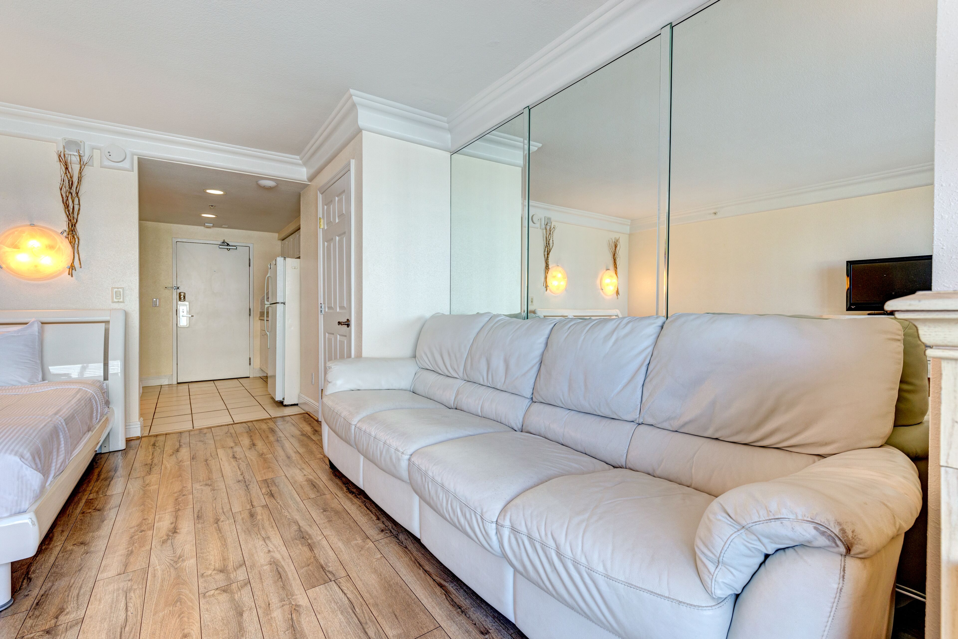 A modern hotel room with a large white leather sofa, a wooden floor, a bed with white bedding, a TV, and a large mirror.