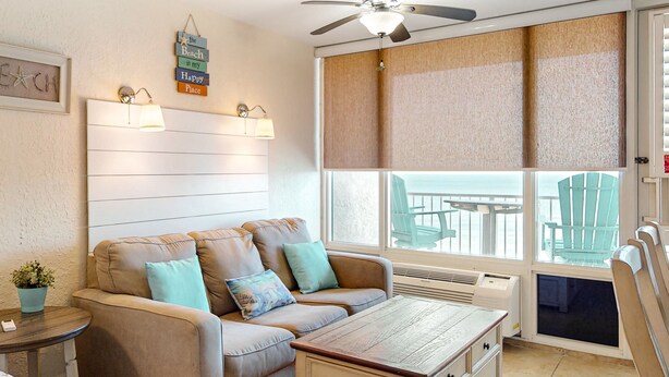 A cozy living room with a beige sofa, a wooden coffee table, and a ceiling fan. There is a large window with a view of outdoor furniture. - Pirates Cove 618 - Sunshine Serenity (Daytona Beach Shores)