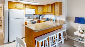 A kitchen with wooden cabinets, a white refrigerator, a microwave, a stove, and a sink. There are three white bar stools and a small white table with a lamp.