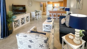 A living room with a sofa, a coffee table, and a TV. There is a dining area with a table and chairs. The kitchen is visible in the background.