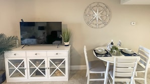 A dining area with a white table set for four, a white cabinet with drawers, a flat-screen TV on a stand, and a decorative wall piece.