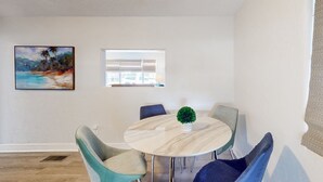 Un comedor con una mesa redonda, cuatro sillas, una planta en maceta y un cuadro con una escena de playa en la pared.