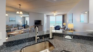 A modern kitchen with granite countertops, stainless steel sink, and a dining area with a table and chairs. - Southpoint 303 (Ponce Inlet)