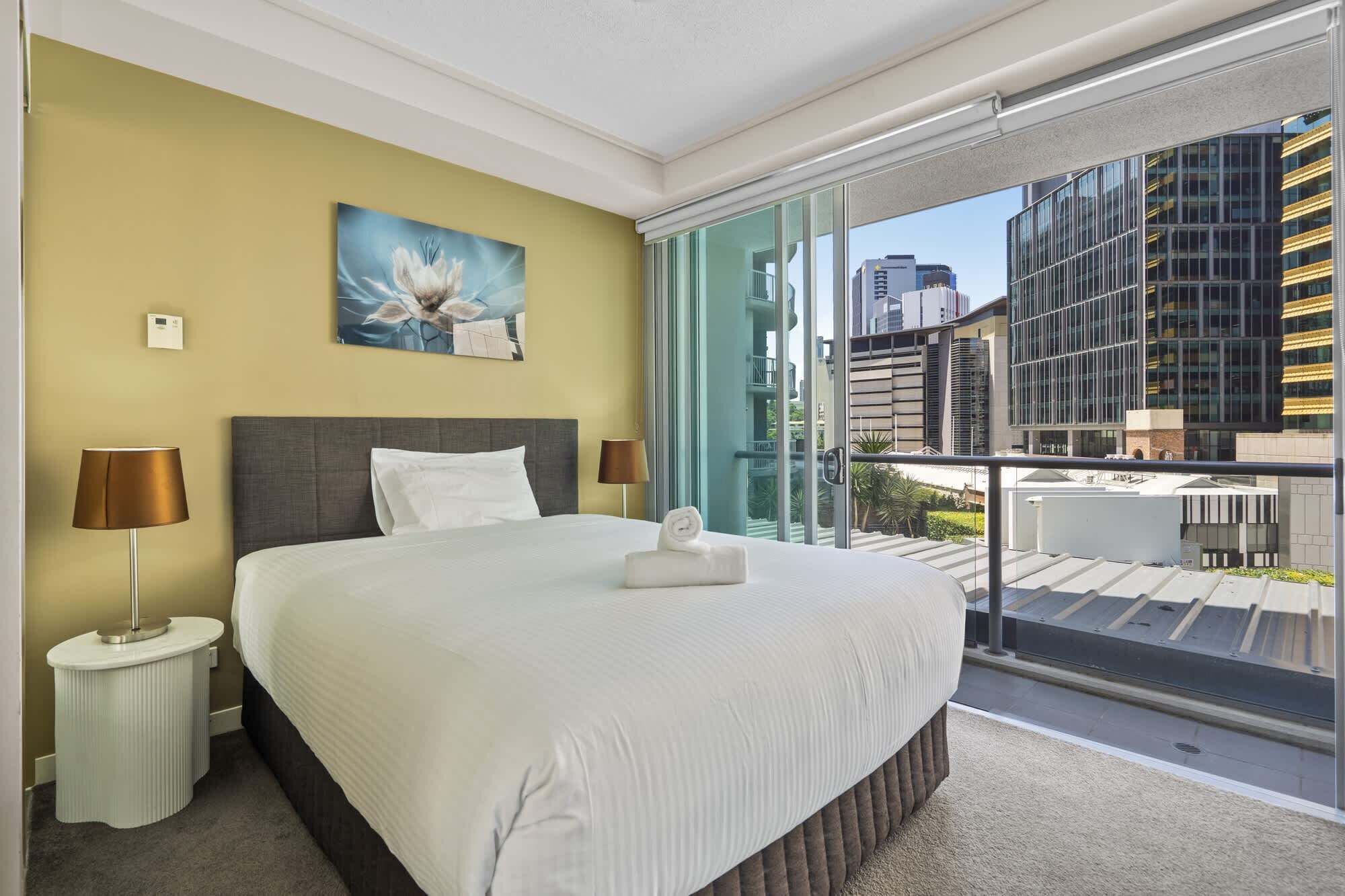 A hotel room with a large bed, a painting on the wall, a window with a city view, and a balcony.
