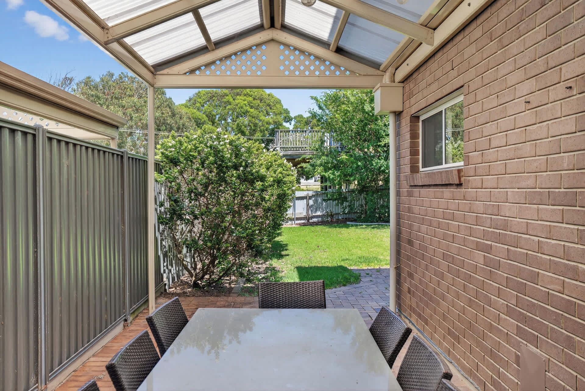 A covered patio with a table and chairs, a brick wall, and a view of a backyard with a tree and a fence.
