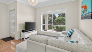 Ein modernes Wohnzimmer mit einem weißen Sofa, einem Flachbildfernseher auf einem weißen Schrank, einem großen Fenster mit Blick ins Grüne und einer Deckenleuchte.