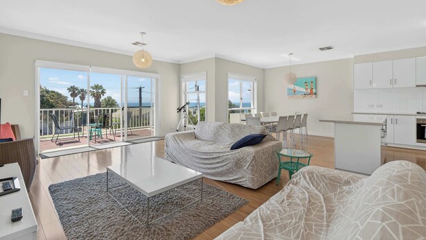 Ein modernes Wohnzimmer mit einer Couch, einem Glastisch und einem Essbereich mit Stühlen. Ein Balkon bietet Blick auf das Meer und Palmen.
