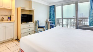 Habitación de hotel con cama, televisor, escritorio con cajones, silla, mesita y balcón con vistas.