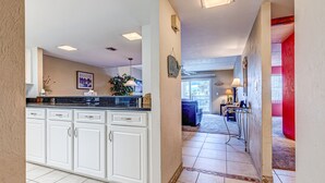 A kitchen with white cabinets and a countertop, a hallway with tiled flooring, and a red accent wall. - Twenty-six Twenty-six 202 - Shores Delight (Daytona Beach)