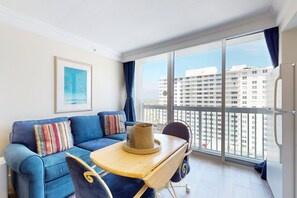 A modern living room with a blue sofa, a small wooden table, and a view of high-rise buildings through large windows. - Daytona Beach Resort 1114 (Daytona Beach)