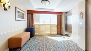 Een hotel lobby met een kamerbreed tapijt, een bank, een prullenbak, een lijst met een afbeelding, een wandlamp en een lift, plus een raam met gordijnen.