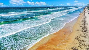 Een strand met golven, een persoon die loopt, en gebouwen op de achtergrond.