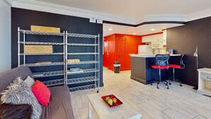 A modern living room with a grey sofa, a metal shelving unit, and a small table with fruit. A kitchen with red accents and a bar area is visible in the background. - Daytona Beach Resort 801 (Daytona Beach)
