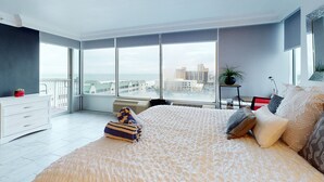 A modern bedroom with a large bed, a white dresser, a small table with a plant, and a view of the city through large windows. - Daytona Beach Resort 801 (Daytona Beach)