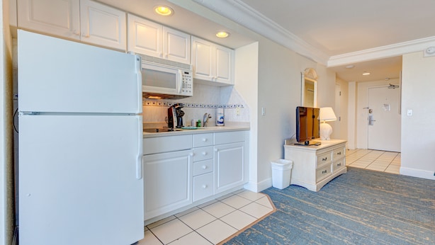 Cocina con armarios blancos, microondas, refrigerador y cafetera. Hay un cubo de basura y un mueble de madera con una lámpara.