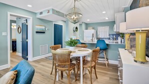 A dining area with a round table, wicker chairs, a chandelier, and a kitchen in the background.