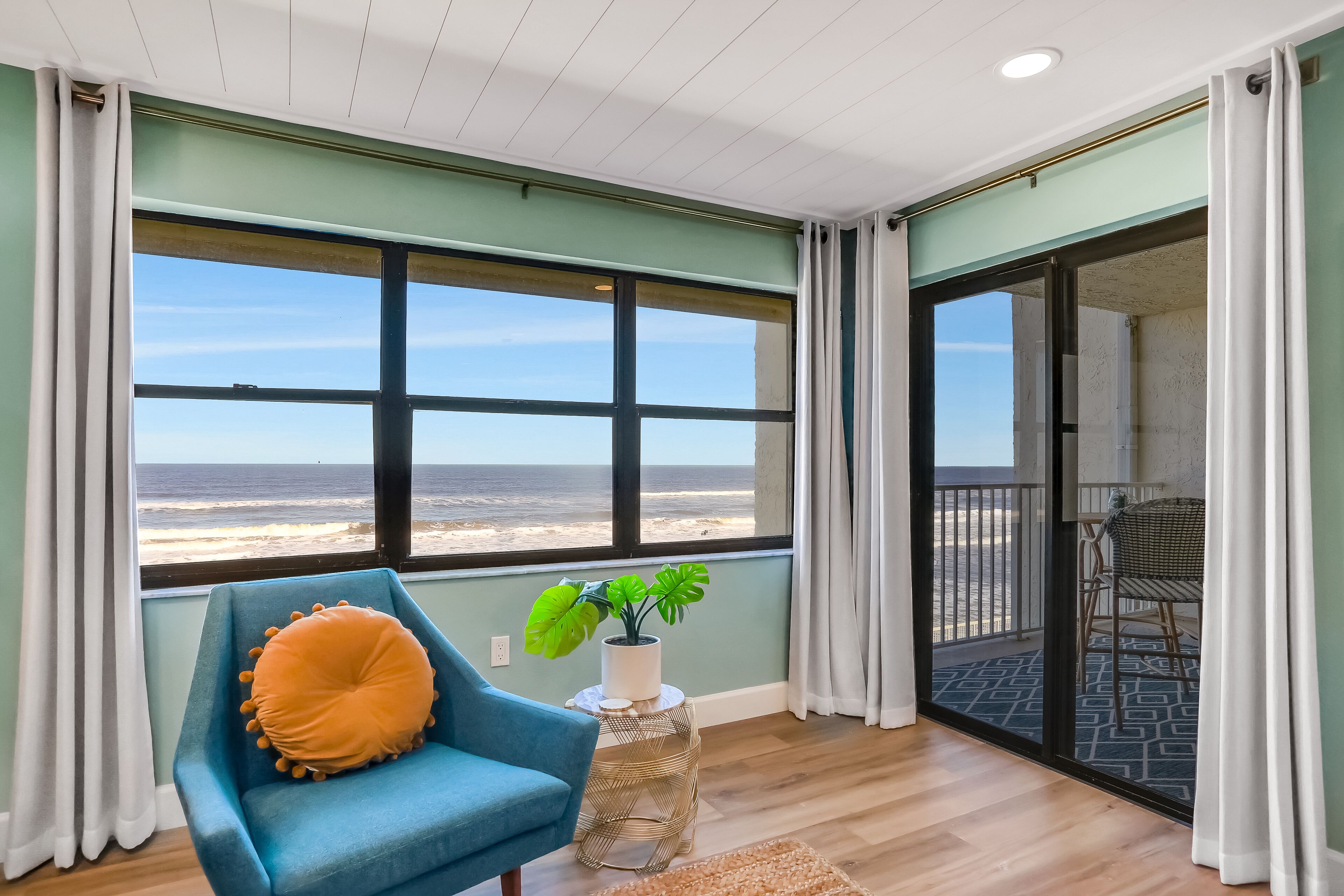 A room with a large window overlooking the beach, a blue armchair, and a small table with a plant.