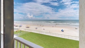 A beach view from a balcony with a grassy area in the foreground and a sandy beach with umbrellas and people in the distance.