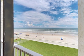 A beach view from a balcony with a grassy area in the foreground and a sandy beach with umbrellas and people in the background.