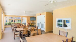 A dining area with wooden tables and chairs, a ceiling fan, and a view of the outside. - Point East 302 -just Beachy (New Smyrna Beach)