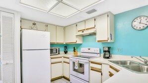 A kitchen with white cabinets, a refrigerator, a microwave, an oven, a sink, and a clock on the wall. - Sea Woods Condo 131 - Coastal Hide Away (New Smyrna Beach)