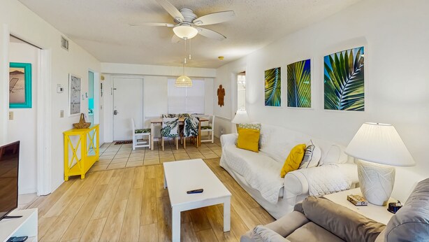 A modern living room with a white sofa, a coffee table, and a dining area with a table and chairs. - Sea Woods Condo 131 - Coastal Hide Away (New Smyrna Beach)