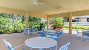 An outdoor patio area with round tables and blue chairs, surrounded by greenery and a white building in the background. - Sea Woods Condo 131 - Coastal Hide Away (New Smyrna Beach)
