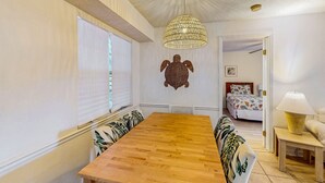 A dining area with a wooden table, patterned cushions, a woven pendant light, and a turtle wall decoration. - Sea Woods Condo 131 - Coastal Hide Away (New Smyrna Beach)