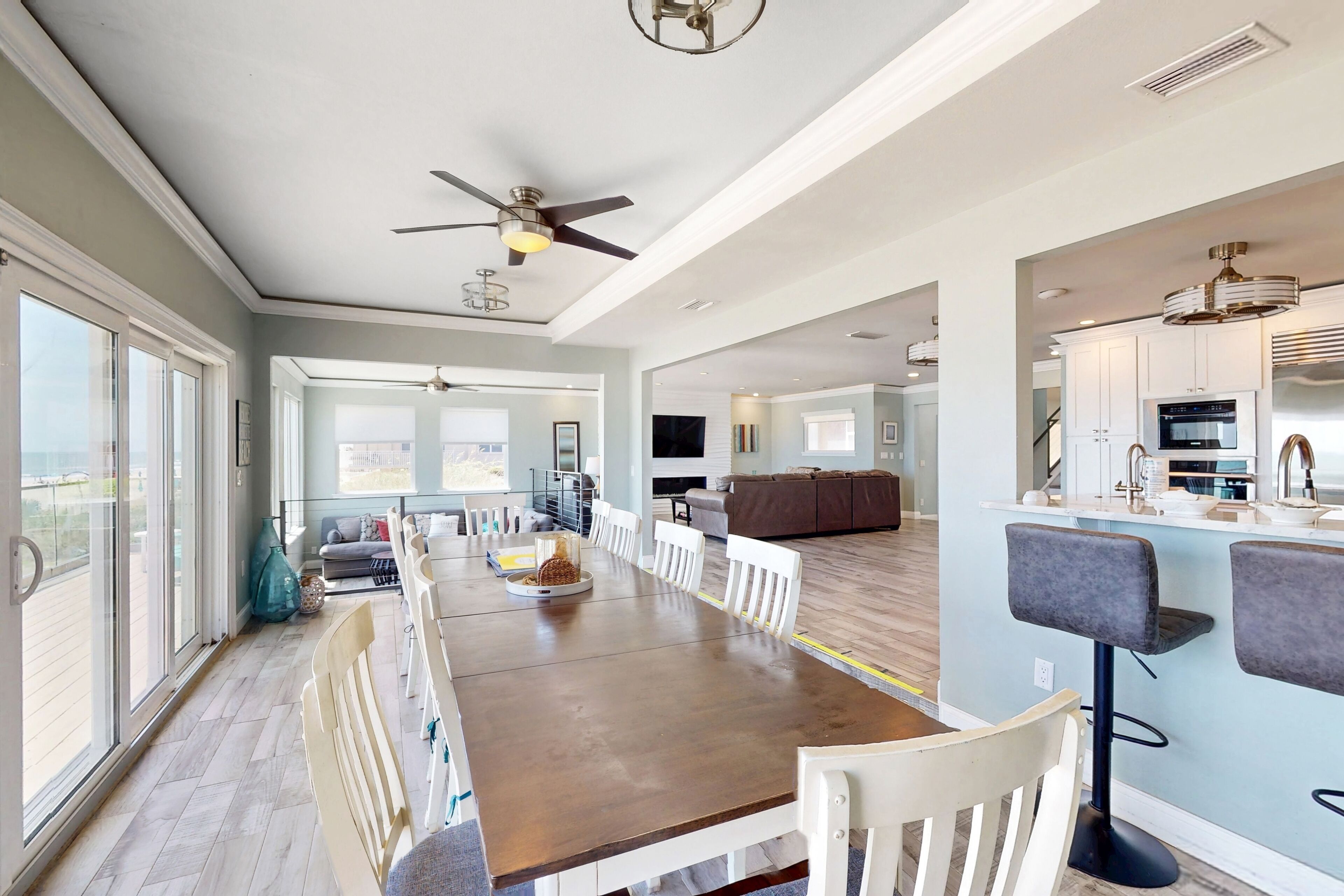 A spacious dining area with a long table and white chairs, a kitchen with modern appliances, and a ceiling fan.