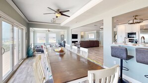 A spacious dining area with a long table and white chairs, a kitchen with modern appliances, and a ceiling fan. - Atlantic Sunrise Home (Daytona Beach Shores)