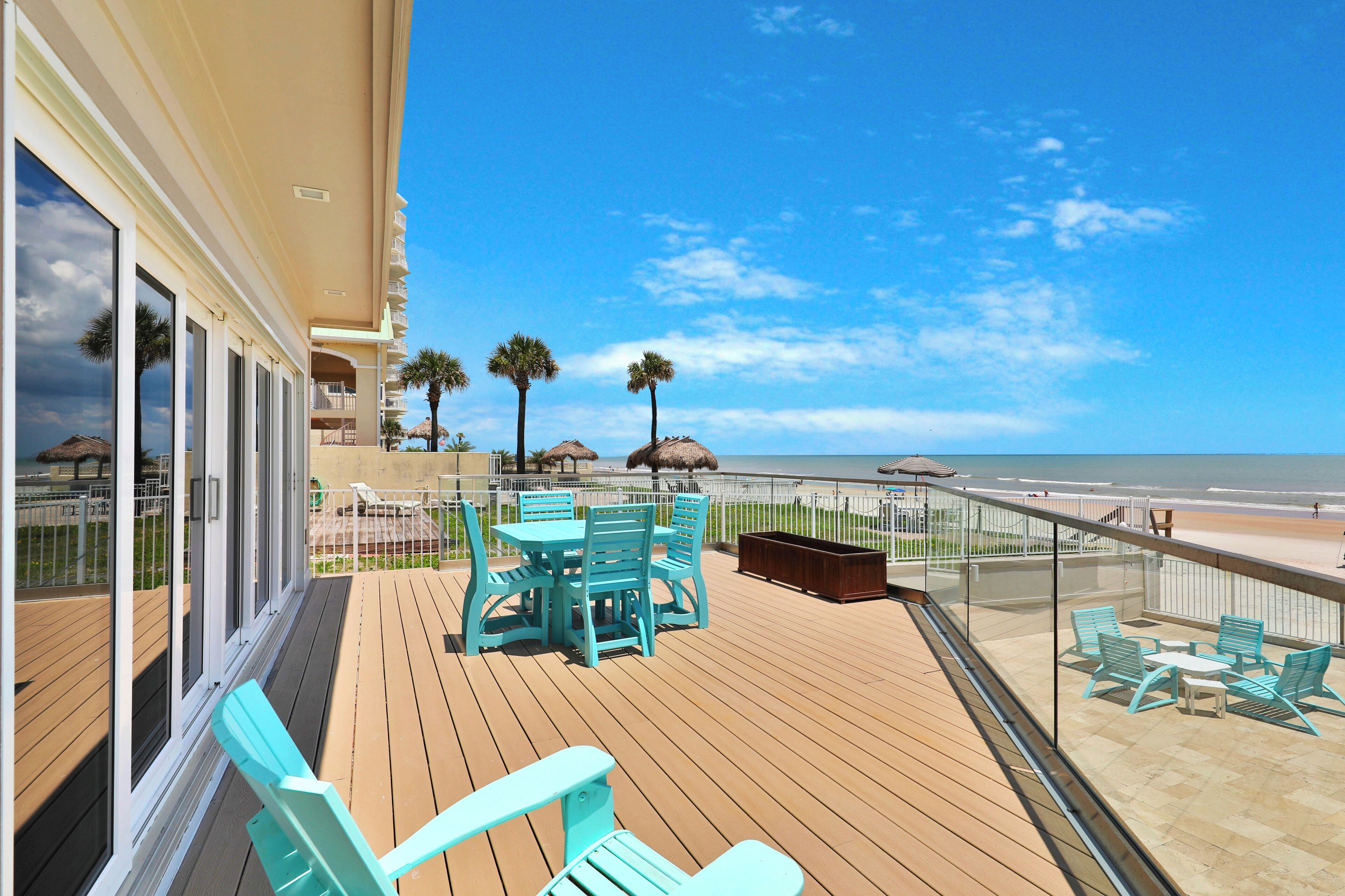 Eine Holzterrasse mit türkisfarbenen Stühlen und einem Tisch, mit Blick auf einen Strand mit Palmen und einem klaren Himmel.