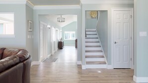 A spacious hallway with a staircase leading to a door, a leather couch, and a chandelier. - Atlantic Sunrise Home (Daytona Beach Shores)