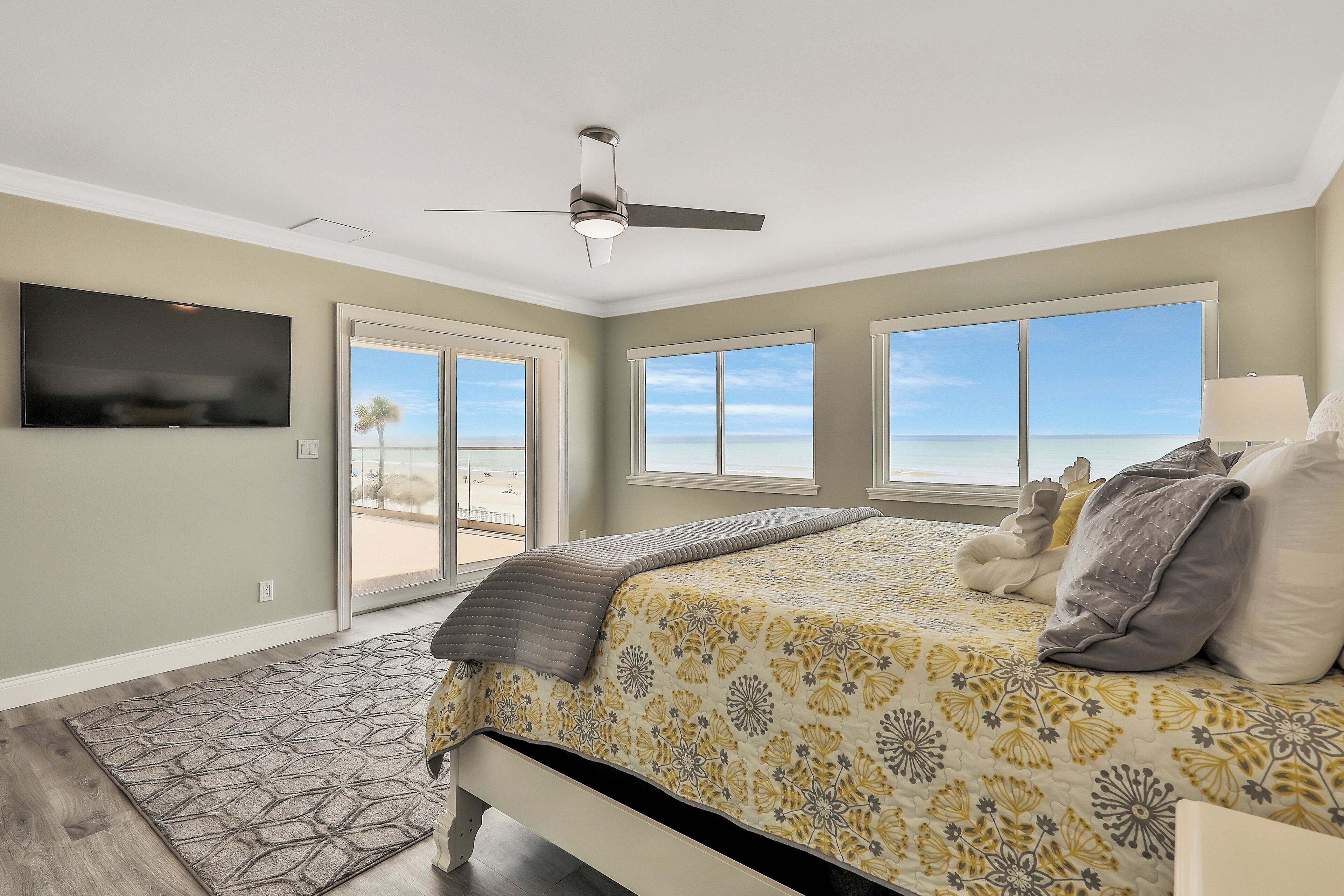 A bedroom with a bed, a ceiling fan, a TV, and large windows with a beach view.