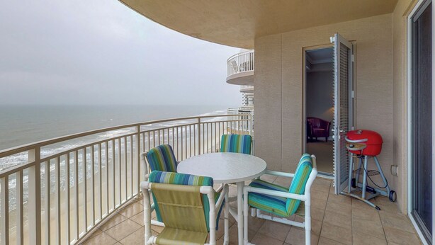 Un balcon avec une table et des chaises blanches, un barbecue rouge et une vue sur l’océan.
