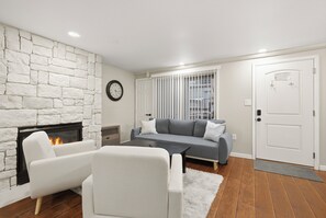 A living room with a stone fireplace, a grey sofa, white armchairs, and a wooden floor. - Park City Walk to a Lifts Edelweiss Haus Condo (Park City)