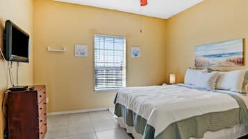 A bedroom with a bed, a dresser, a television mounted on the wall, a ceiling fan, and a window with blinds.