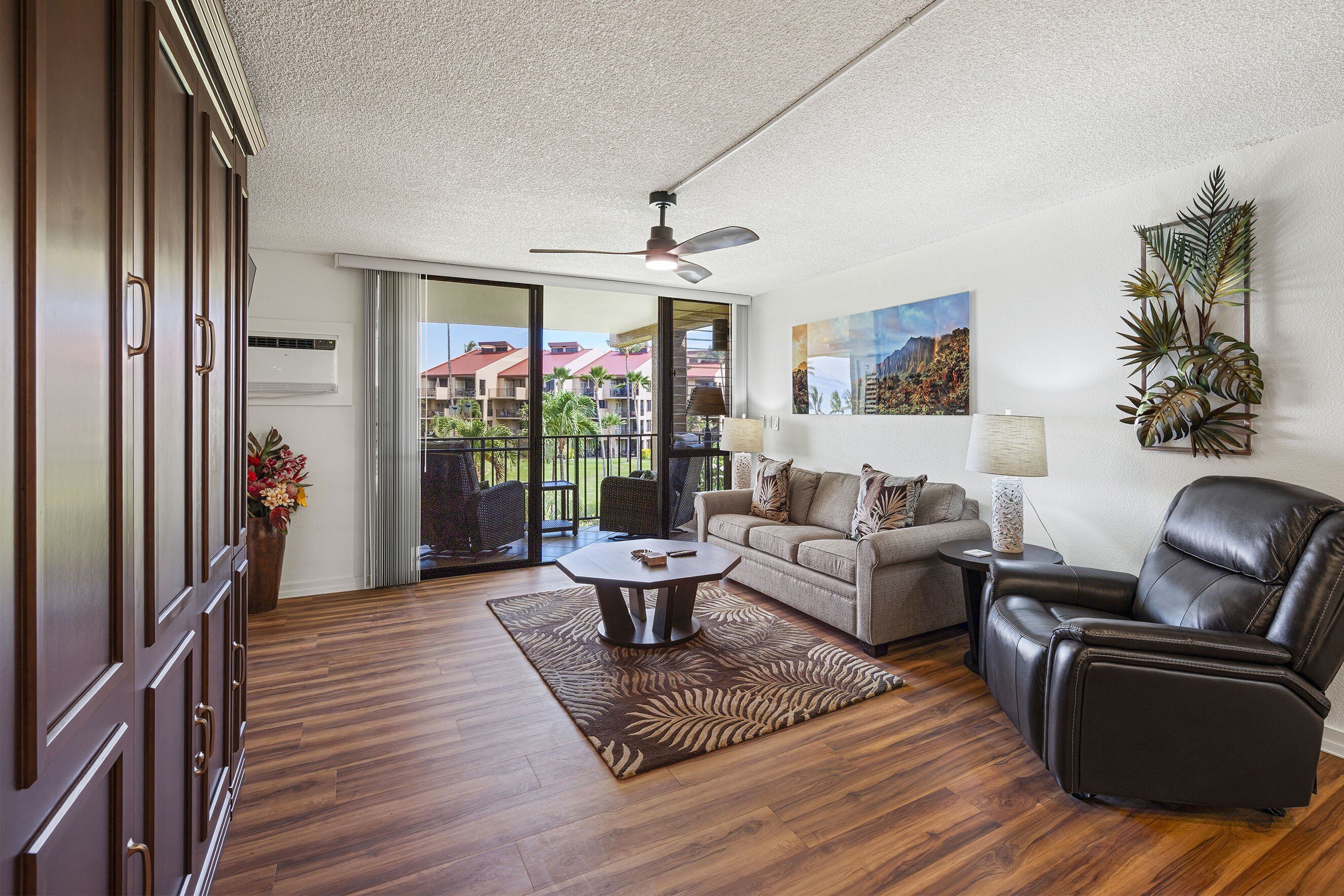 A spacious living room with a wooden floor, a ceiling fan, a sofa, a recliner, a coffee table, and a large window offering a view of buildings and greenery.