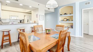 A dining area with a wooden table and chairs, a kitchen in the background, and a bookshelf with various items. - Oceanwalk 16-305 (New Smyrna Beach)