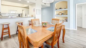 A dining area with a wooden table and chairs, a kitchen in the background, and a bookshelf with various items.