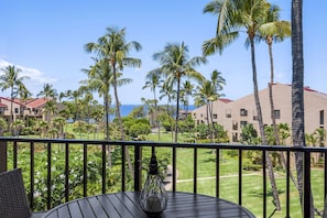 A balcony with a view of a well-maintained lawn, palm trees, and a building with a red roof. - Kamaole Sands 5-414 (Kihei)