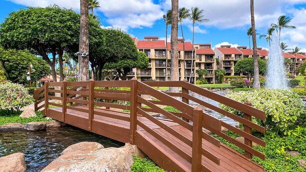 A wooden bridge over a small stream, surrounded by lush greenery and palm trees, leading to a building with multiple balconies and a red roof. - Kamaole Sands 5-414 (Kihei)