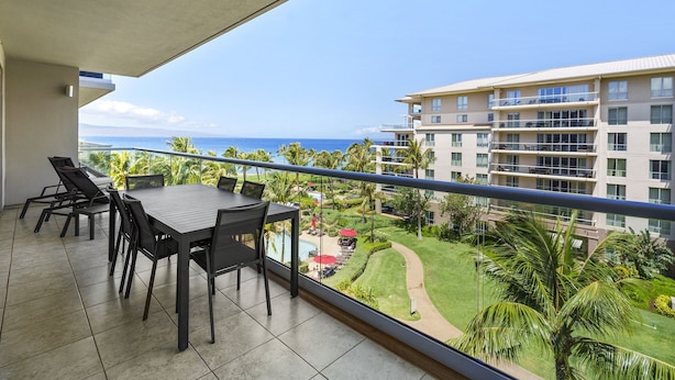 A balcony with a table and chairs, overlooking a pool and lush greenery, with a view of a hotel building and the ocean. - Honua Kai Konea 505 (Kaanapali)
