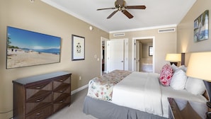A bedroom with a bed, a dresser, a ceiling fan, a beach-themed painting, and a door leading to another room. - Honua Kai Konea 505 (Kaanapali)