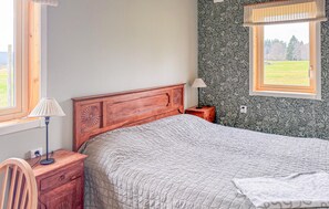 A bedroom with a wooden headboard, a single bed with a quilted blanket, a bedside table with a lamp, a nightstand, and a window with a view of a green field. - 2 bedroom stunning home in Sandhem (Sandhem)