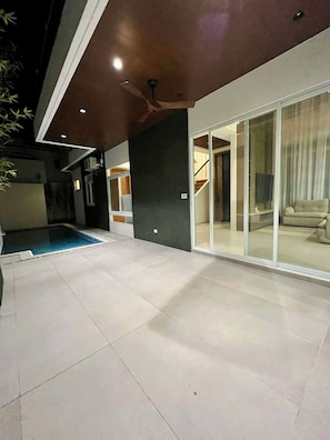 A modern outdoor space with a swimming pool, a covered patio, and a glass door leading to an interior area.
