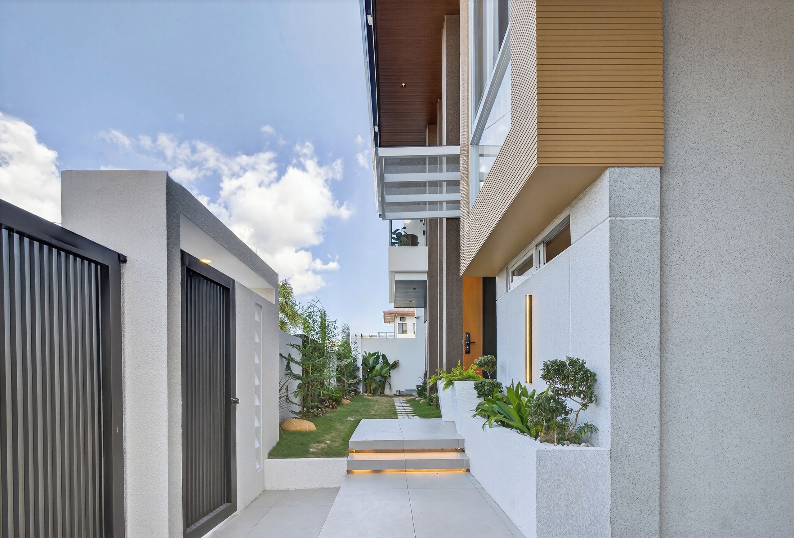 A modern residential building with a narrow pathway leading to the entrance, surrounded by greenery and a clear sky.