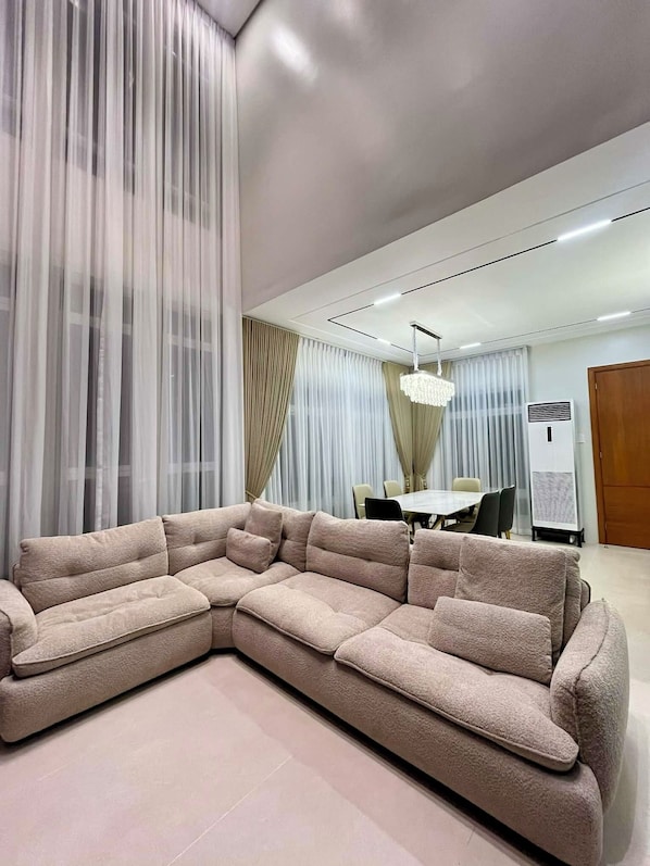 A modern living room with a large sectional sofa, a dining area with a table and chairs, and a large window with sheer curtains.