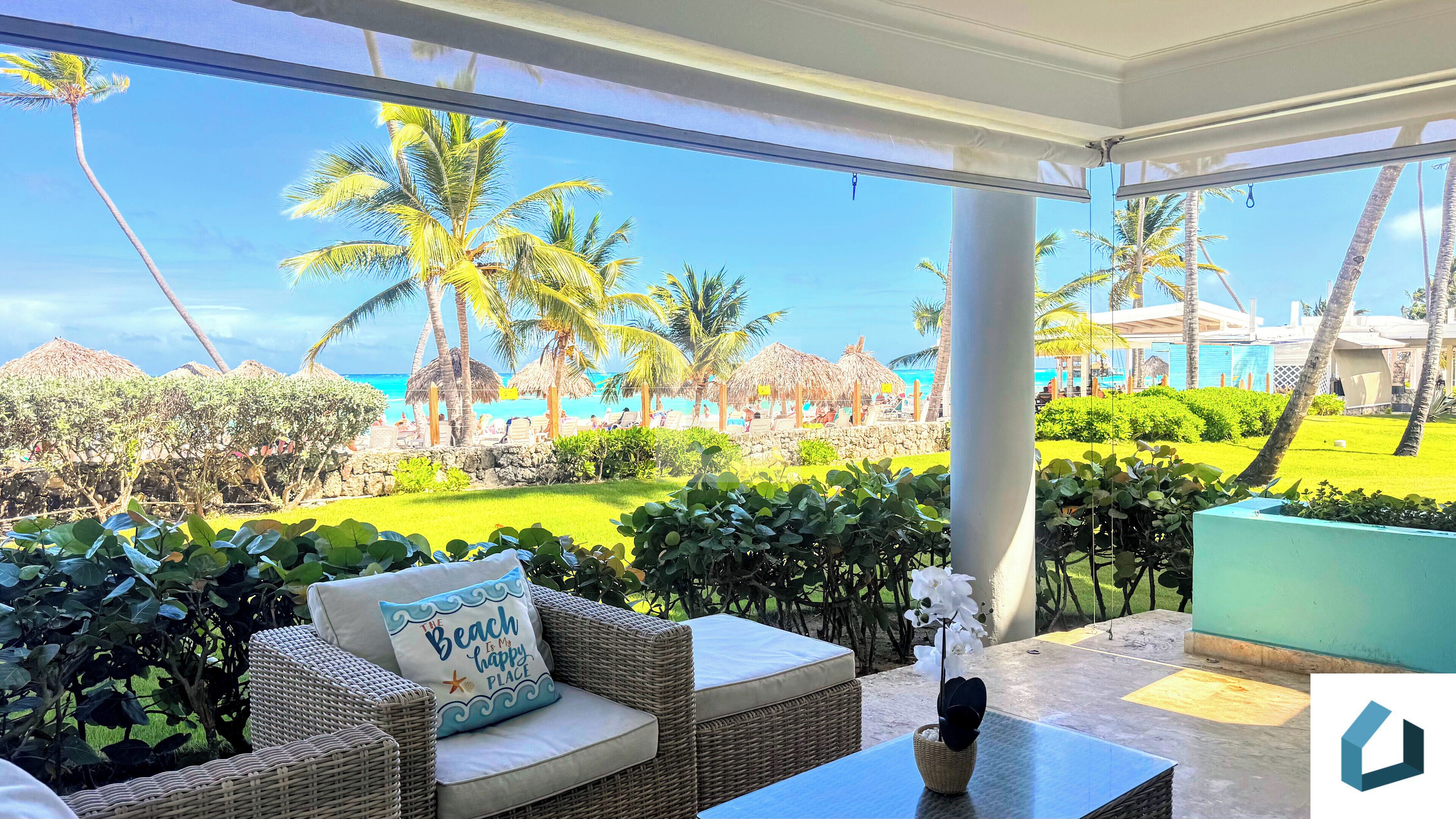 A balcony with wicker furniture, a cushion with a beach-themed pillow, and a view of a beach with palm trees and a pool.