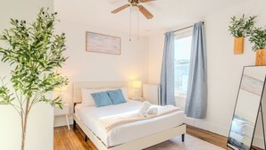 A bedroom with a bed, a ceiling fan, a bedside table, a nightstand, a window with curtains, a potted plant, and a mirror. - Cozy 3Bed only 4min from tufts-10min from downtown Boston (Medford)